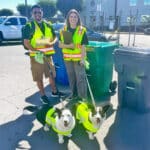 Alameda Post - Next to three bins, two trash inspectors smile at the camera. They hold clipboards and are accompanied by two dogs. All four of them wear safety vests.
