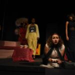 Alameda Post - actors in Antigone 3021 perform on a dimly lit set. One actress kneels close to the camera and gestures with her hands while the others look on