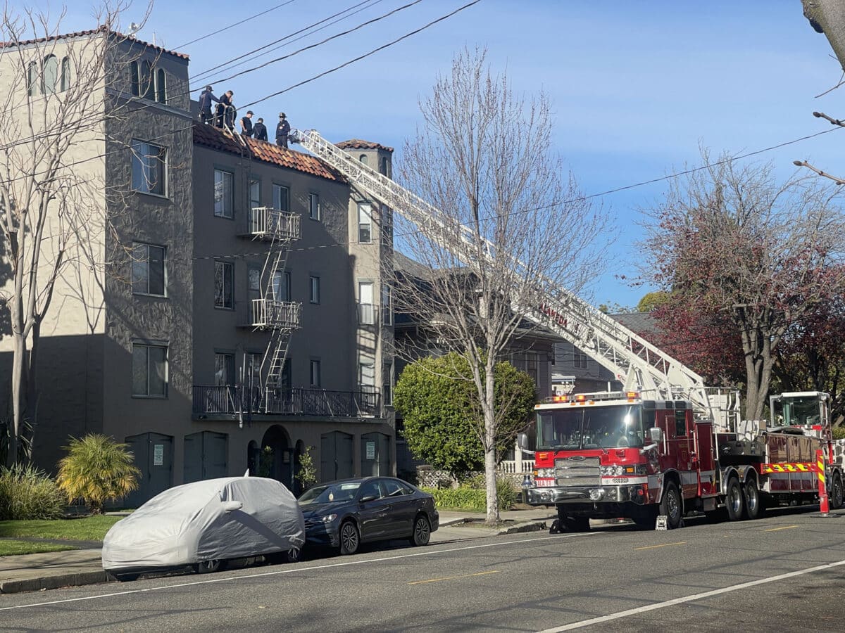Alameda Post - on top of a four or so story apartment building, firefighters stand around. A firetruck is parked next to the building, and a ladder goes to the top.