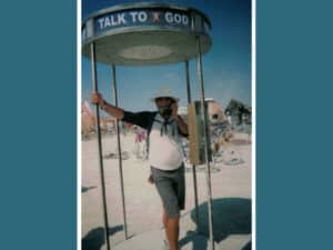 Alameda Post - a photo of Joseph Bryan "Joe" Devine. He is standing in a type telephone booth, which says "Talk to God."