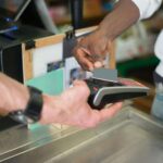 Alameda Post - a cashier holds out a card reader, and a customer taps their card
