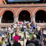 Alameda Post - before the People's March in Alameda, community members gather at City Hall holding signs while someone speaks into a microphone