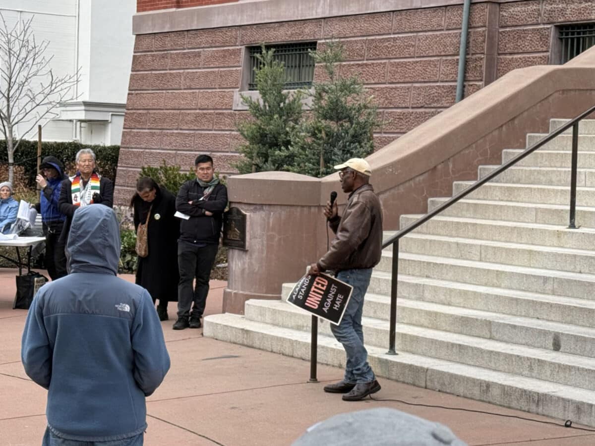 Alameda Post - a man holds a microphone in front of the steps of City Hall and speaks to a crowd