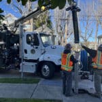 Alameda Post - two workers in bright orange vests maneuver a large pipe connected to a truck into the ground under a grate