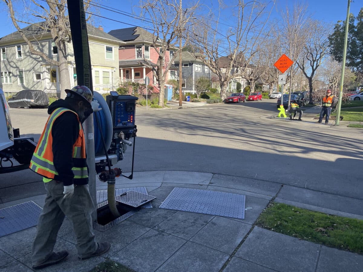 Alameda Post - a worker in an orange utility vest looks into the ground under a grate while machinery is attached to it from a truck