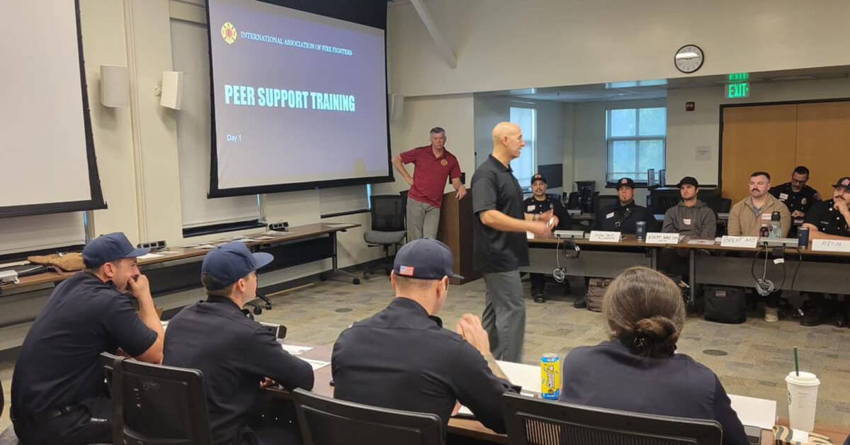 Alameda Post - firefighters sit in a classroom while a man stands up to give a presentation. On a projector, a slide says "Peer Support Training"