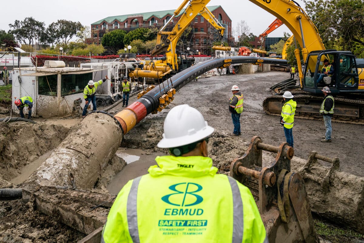 Alameda Post - a crew of EBMUD workers in brightly colored gear install a huge pipe into the ground