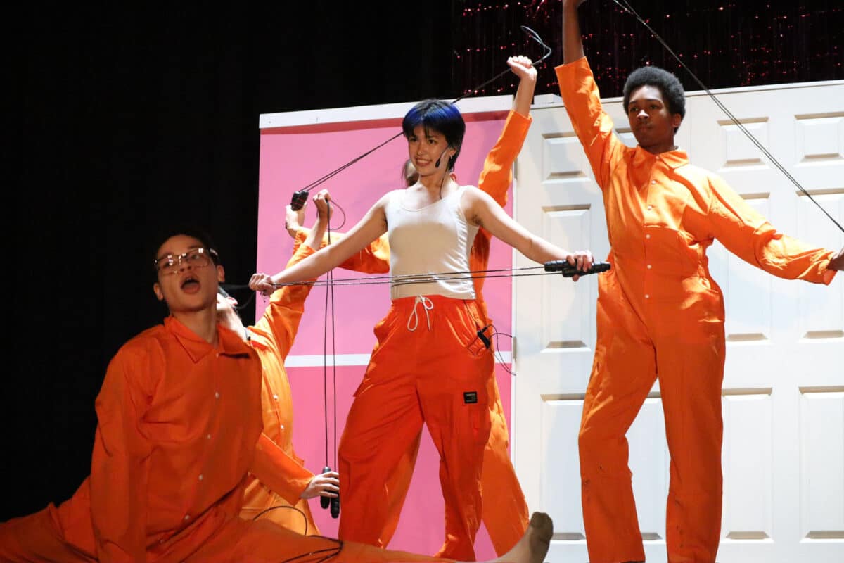 Alameda Post - an ensemble in prison jumpsuit orange strike a pose while singing