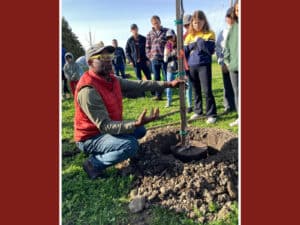 Alameda Post - a man crouches on the ground next to a sapling tree and talks to a group of people