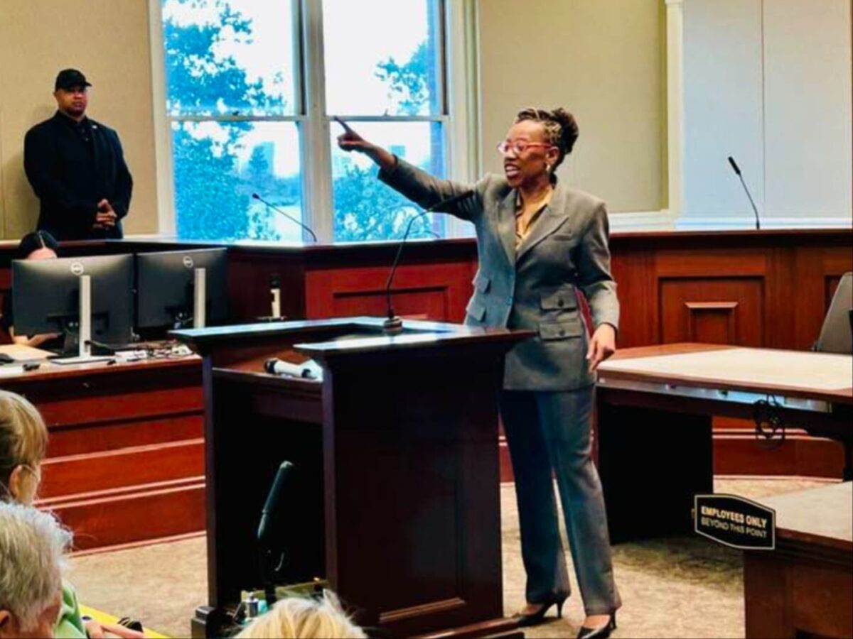 Alameda Post - Lateefah Simon stands in a suit and heels at the front of Town Hall. She is speaking animatedly and points to the back of the room