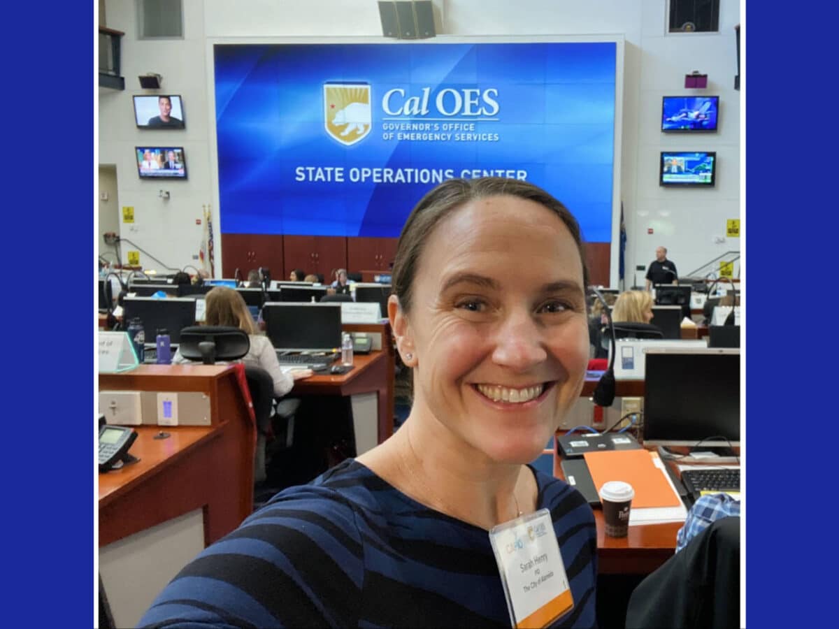 Alameda Post - Sarah Henry takes a selfie while in a large conference room in front of a projector screen that says CAL OES State Operations Center