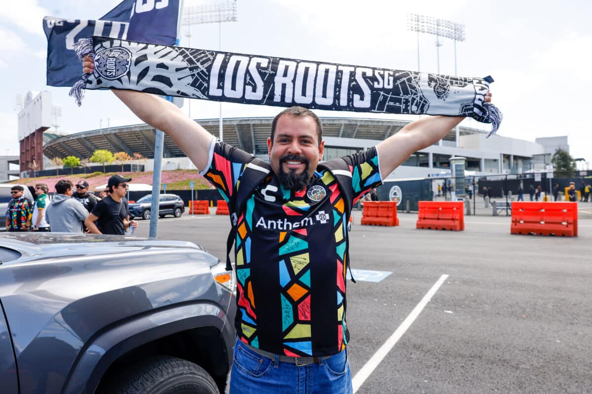 Alameda Post - an Oakland Roots fan wearing a jersey holds up a scarf that says "Los Roots" in front of the Colosseum before the match versus Orange County SC