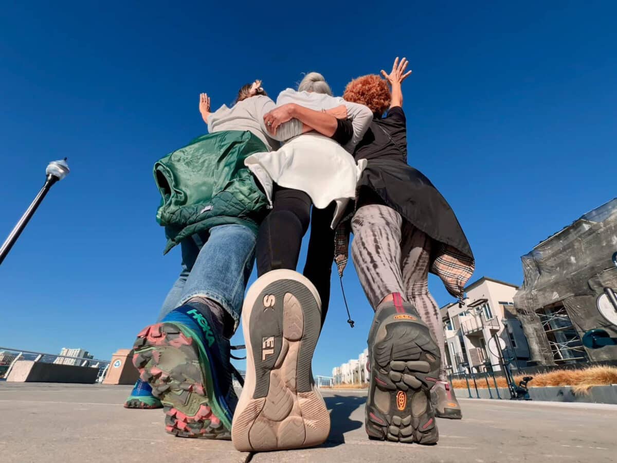 Alameda Post - a group of three women in walking shoes strike a pose. The camera is set behind them, so their shoes and backs are in frame.