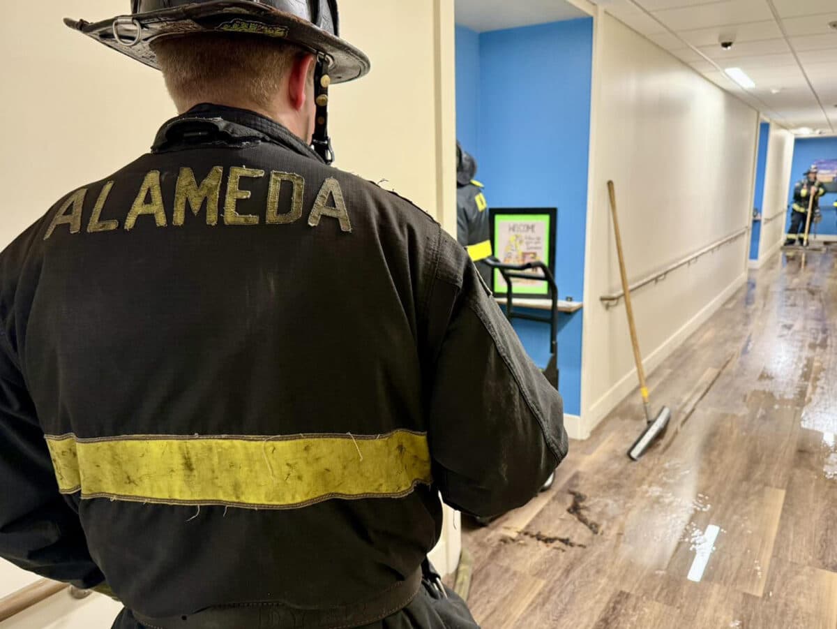 Alameda Post - an Alameda Fire Department firefighter walks through a brightly lit hallway that is covered with water.