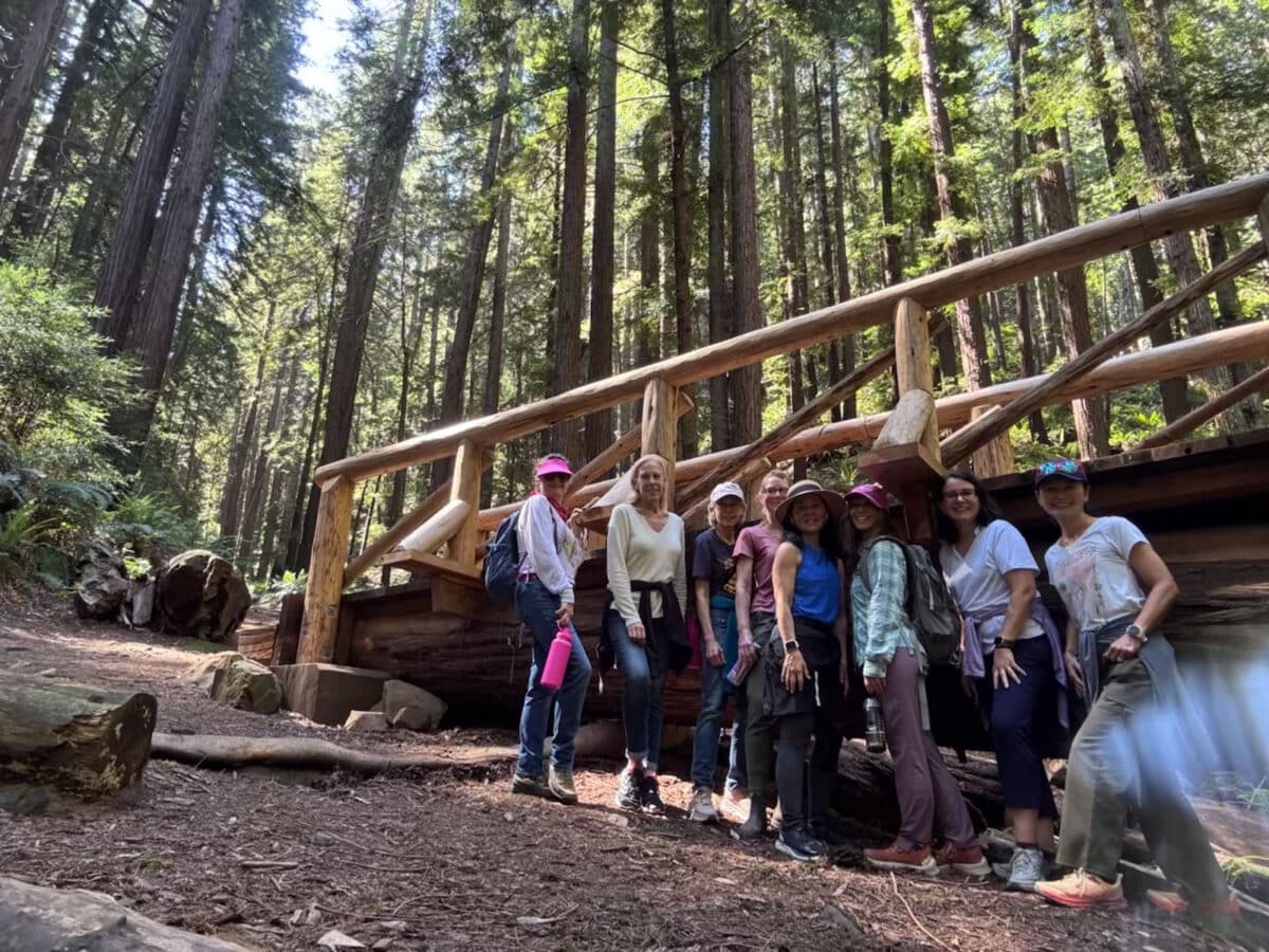 Alameda Post - eight women in hiking gear take a break by a wooden structure amongst a redwood forest