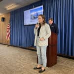 Alameda Post - A man stands behind a podium, and a woman holds a microphone and speaks into it while a slideshow shows behind her