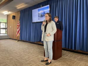 Alameda Post - A man stands behind a podium, and a woman holds a microphone and speaks into it while a slideshow shows behind her