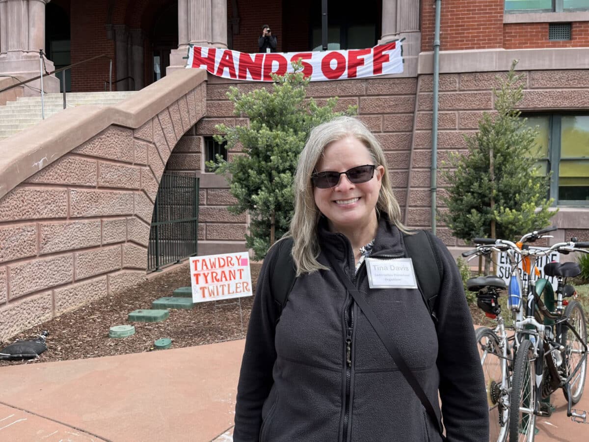 Alameda Post - a woman, Tina Davis, smiles in front of City Hall. A banner behind her says "Hands Off"
