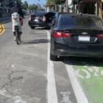 Alameda Post - a parent with a small child on their bike swerves around several parked cars in the bike lane.