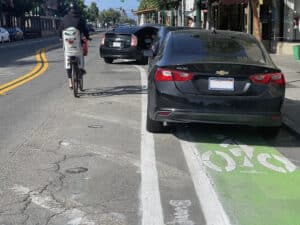 Alameda Post - a parent with a small child on their bike swerves around several parked cars in the bike lane.