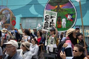 Alameda Post - participants in the Right of Return Pilgrimage gather. They look towards someone or something out of frame, raising their hands balled into fists, and holding sights that say things like "Interfaith Pilgrimage for Peace"