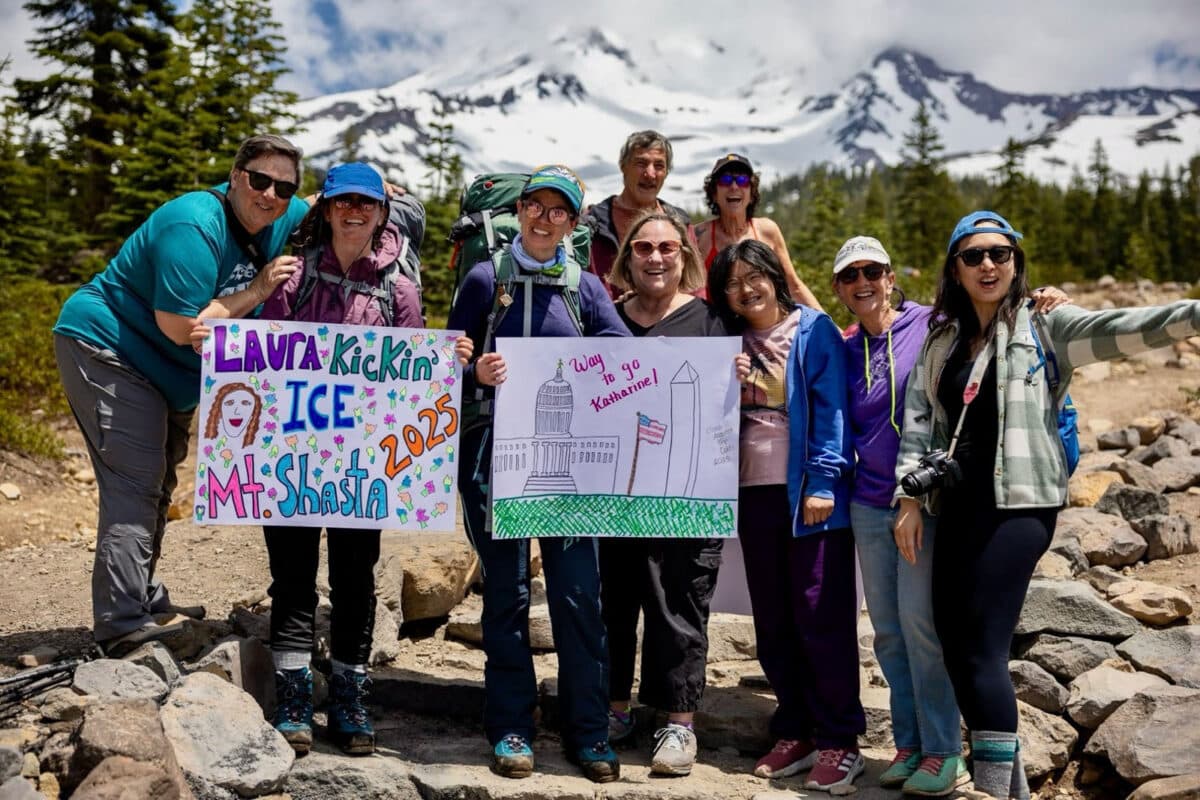 Alameda Post - a group of women stand at the base of mount Shasta holding signs that says things like "Way to go Katharine" and "Laura Kickin' Ice Mt. Shasta 2025"
