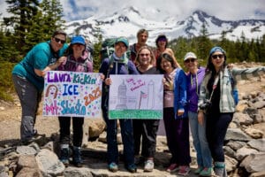 Alameda Post - a group of women stand at the base of mount Shasta holding signs that says things like "Way to go Katharine" and "Laura Kickin' Ice Mt. Shasta 2025"