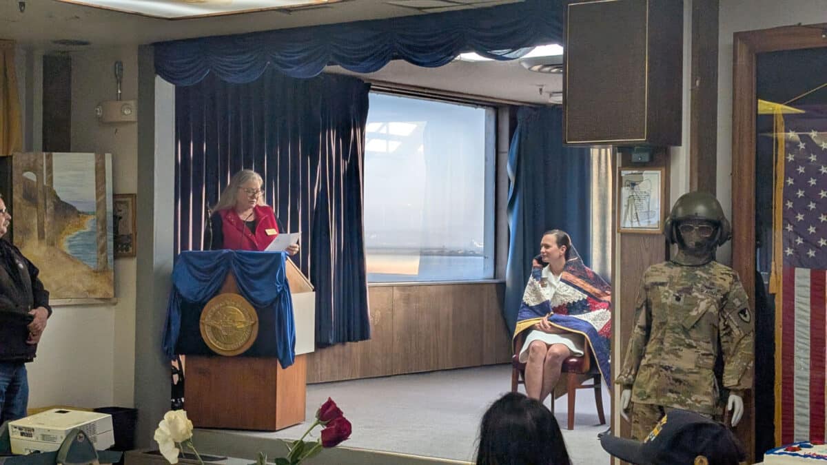 Alameda Post - during a ceremony, one woman reads from a pulpit while another woman is seated onstage and wrapped in a quilt
