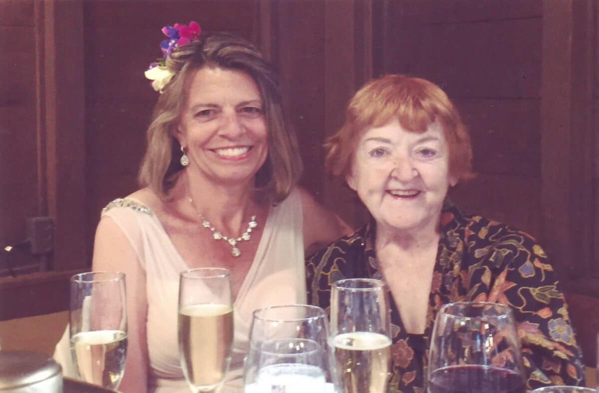 Alameda Post - two women, one in a bridal gown, sit at a table with glasses of champagne