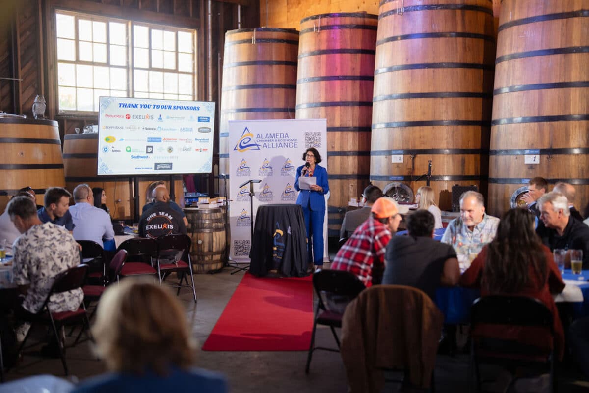 Alameda Post - in front of large brewery containers at a venue, Mayor Ashcraft wears a royal blue suit and holds a microphone. She talks to a crowd seated at tables at the Innovation Island Excellence Awards