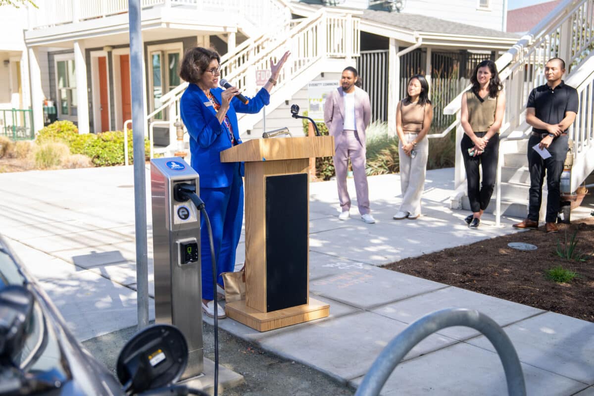 Alameda Post - a woman in a bright blue suit, Alameda's current mayor, stands at a podium and speaks into a microphone. She is outdoors on a walkway and standing next to a new EV charger. A group of four people stand off to the side.