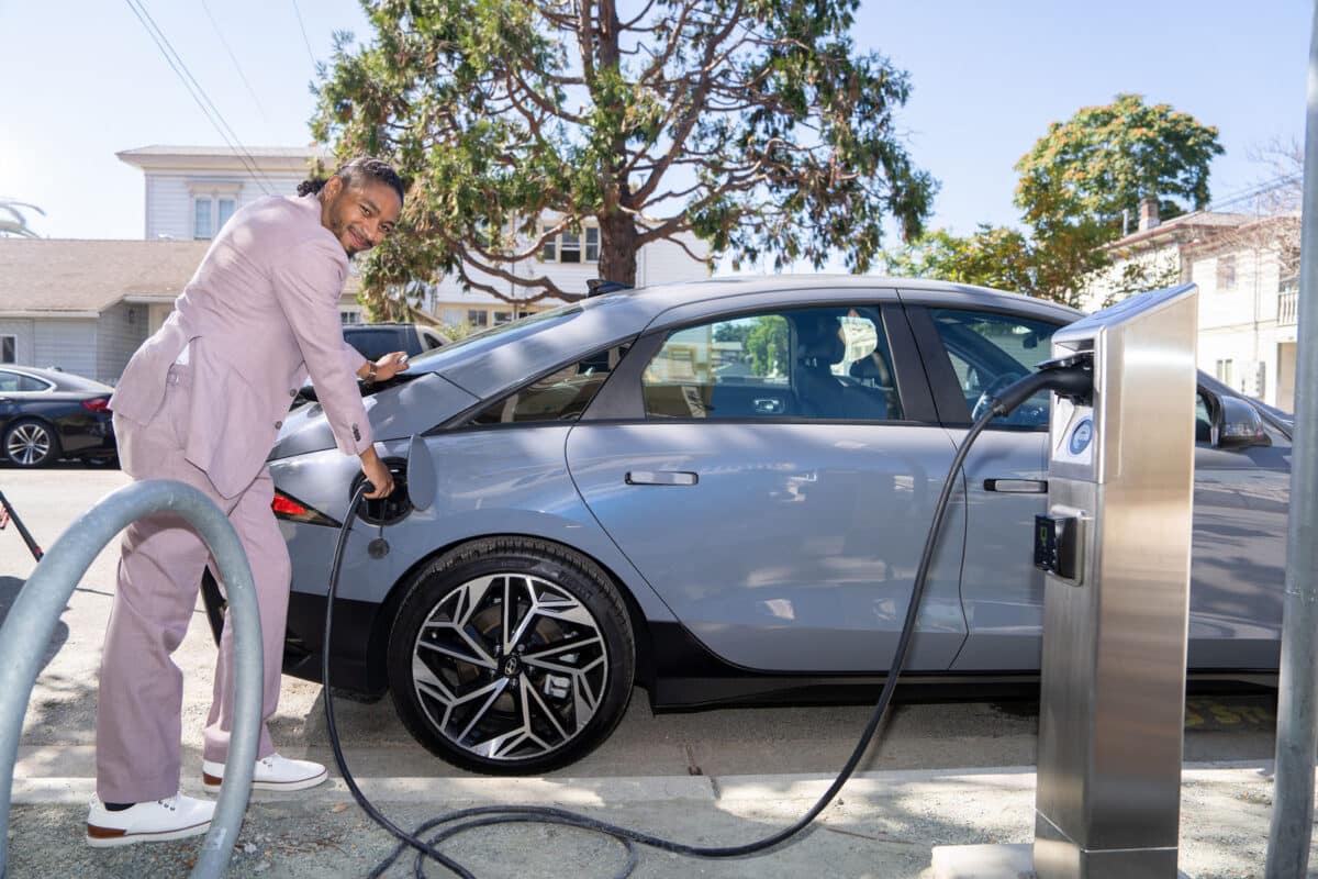 Alameda Post - A man in a light blue suit smiles at the camera and inserts a charger into an electric vehicle.