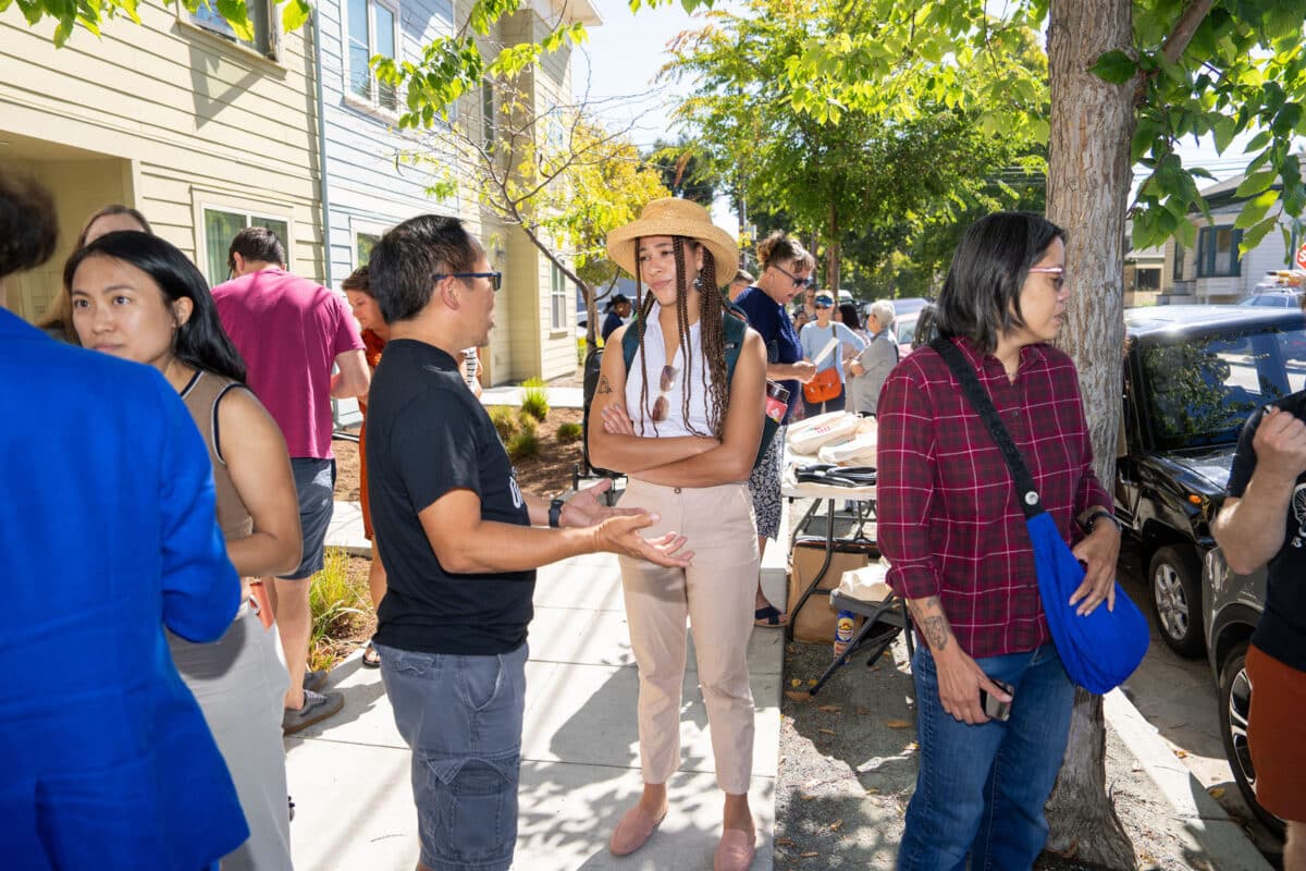 Alameda Post - People talk and mingle on the sidewalk at an EV charger ribbon cutting event.