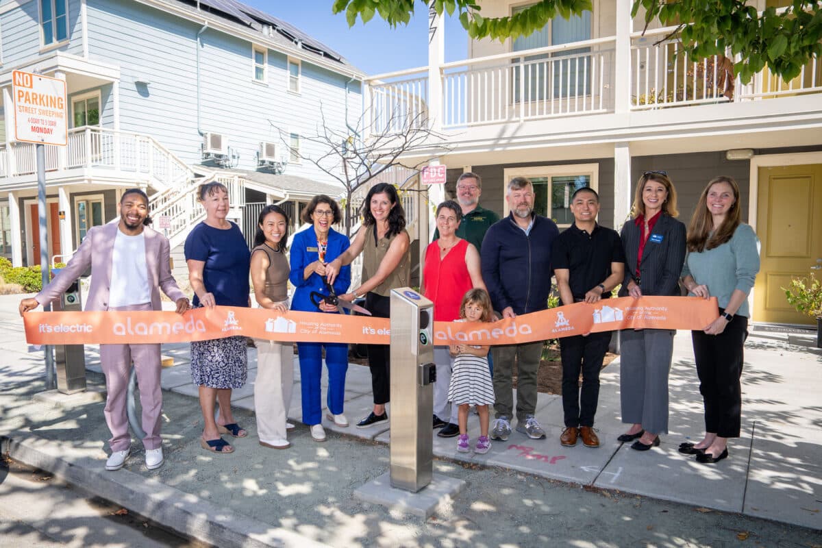 Alameda Post - 11 adults and one child gather around a long ribbon and smile at the camera. They mime a ribbon cutting of a new EV charger next to the sidewalk.