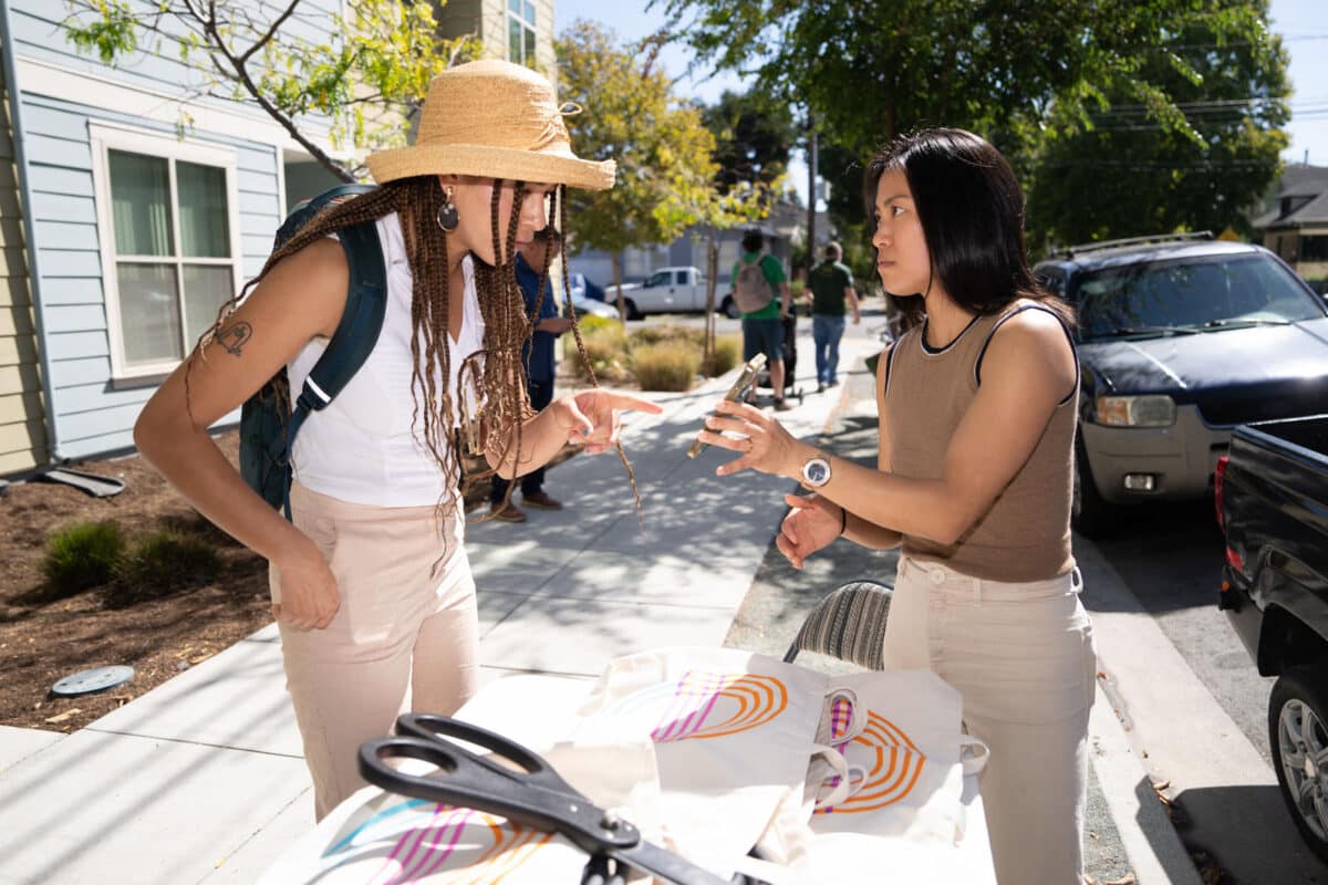 Alameda Post - at an information booth on the sidewalk, one person holds a phone and explains something to another person.
