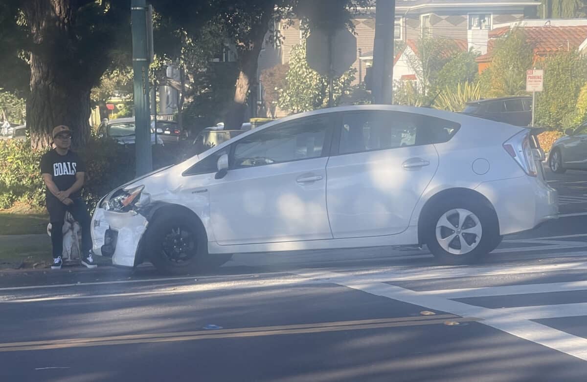Alameda Post - a person sits next to a white prius with an obvious broken and dented front bumper