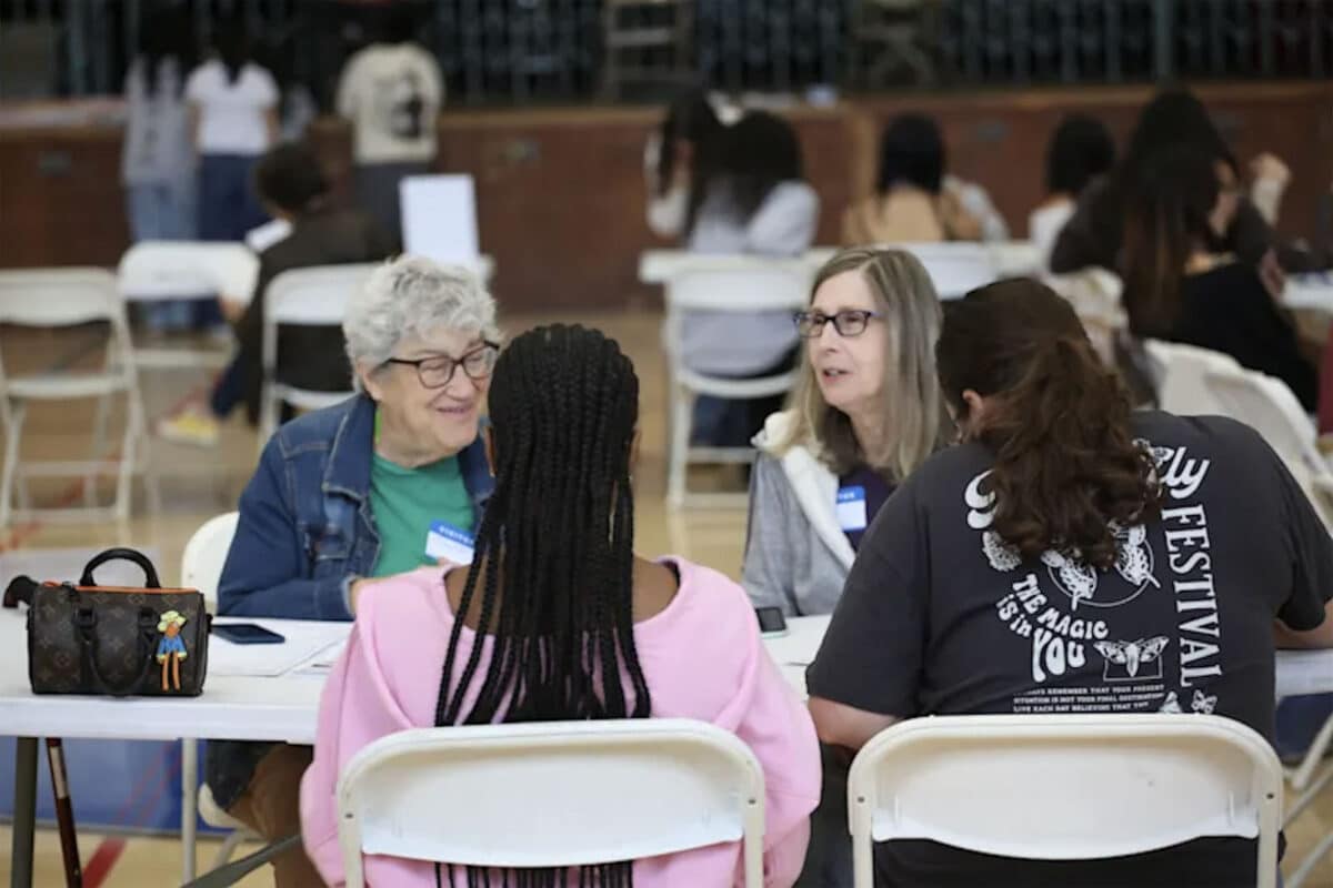 Alameda Post - two women with name tags sit at a table with two high school students. They are smiling and appearing to be doing paperwork with the high schoolers.