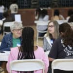 Alameda Post - two women with name tags sit at a table with two high school students. They are smiling and appearing to be doing paperwork with the high schoolers.