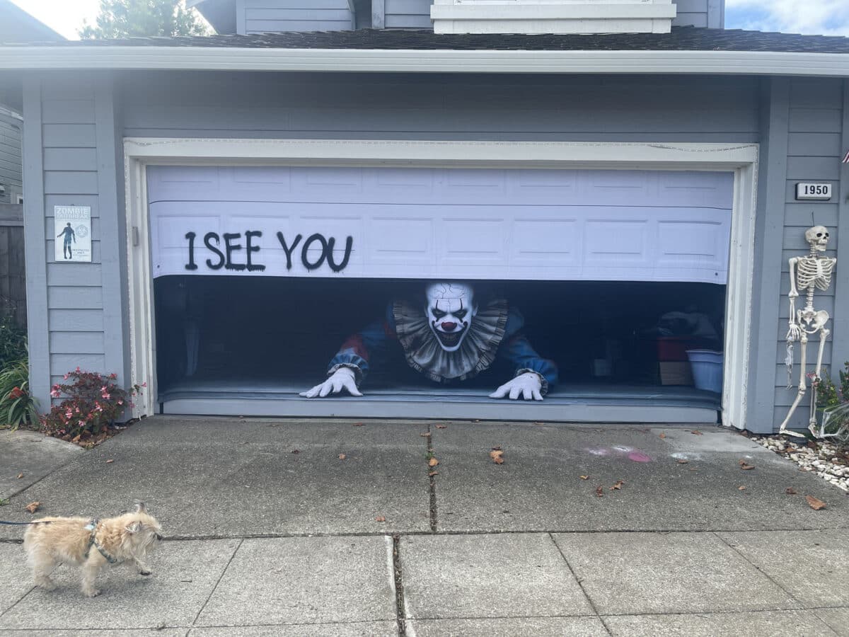 Alameda Post - A decorative screen or tarp across a garage door. The screen is very realistic to look just like a garage door with a scary clown crawling out from a partially open garage door. Scrawled letters say "I see you"