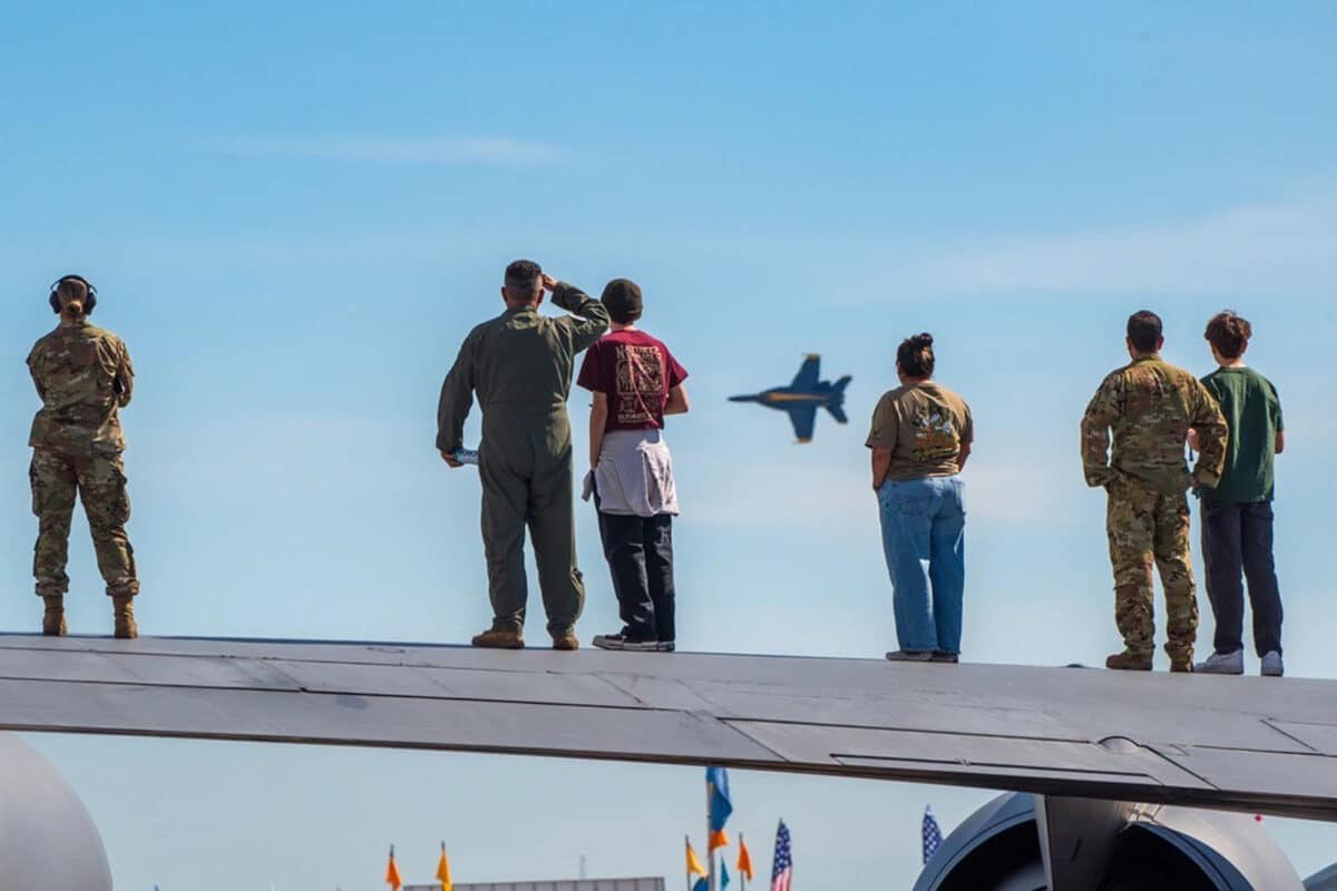 Alameda Post - Military personal stand on the wings of a plane and salute a Blue Angel flying above them.