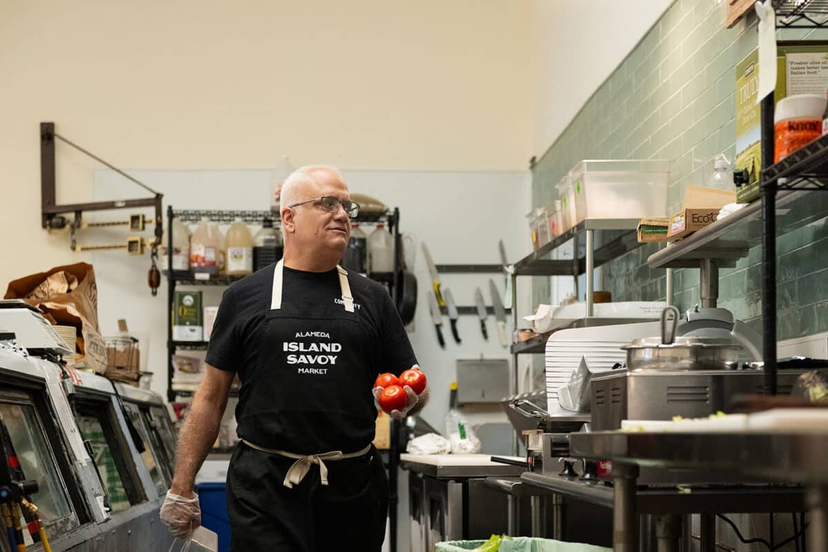 Alameda Post - Mike Stevens walks through the deli of Island Savoy Market holding tomatoes. 
