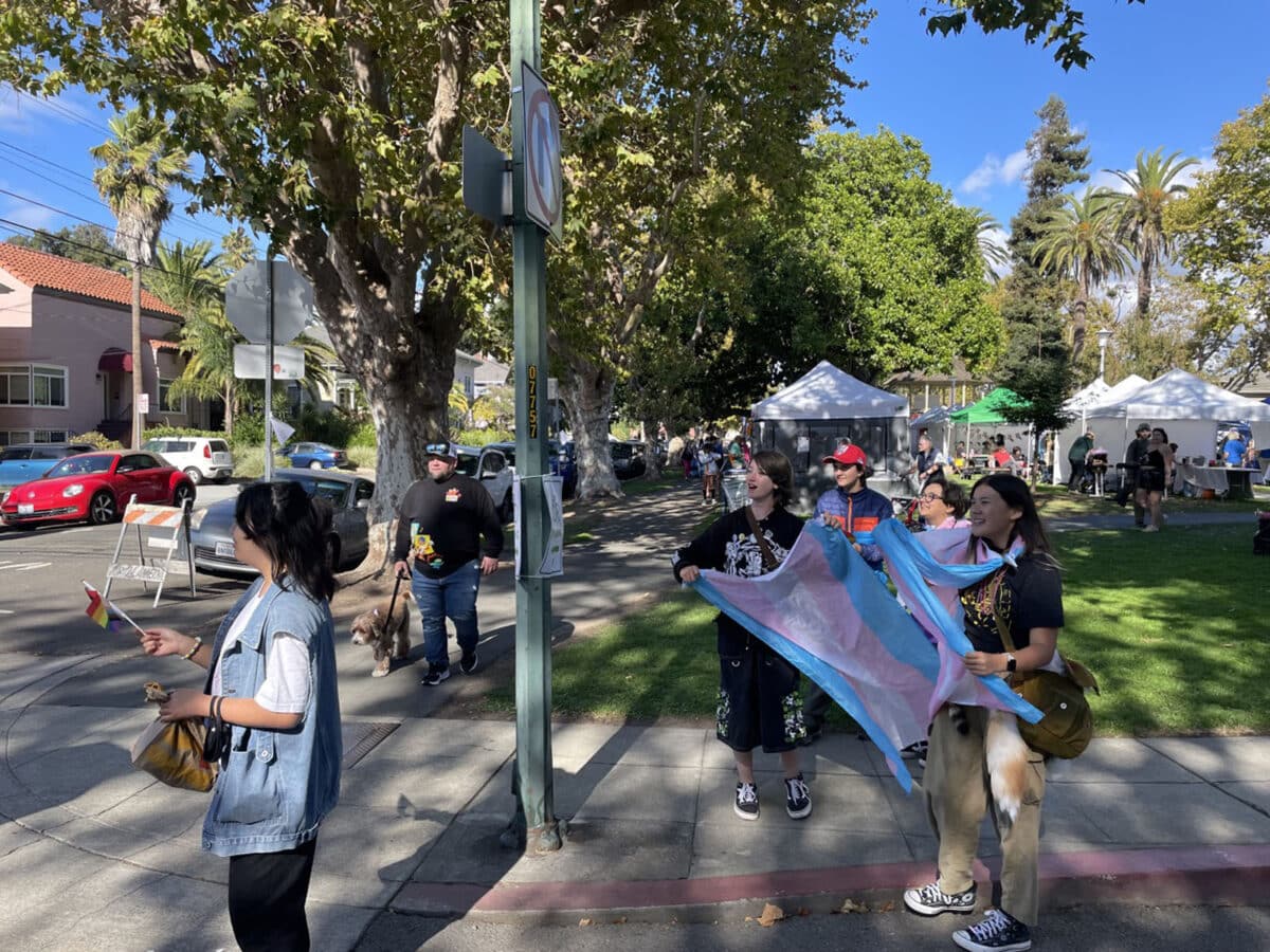 Alameda Post - A group of young, perhaps high school aged, attendees smile and wave pride flags in the same direction towards a protestor who is out of frame.