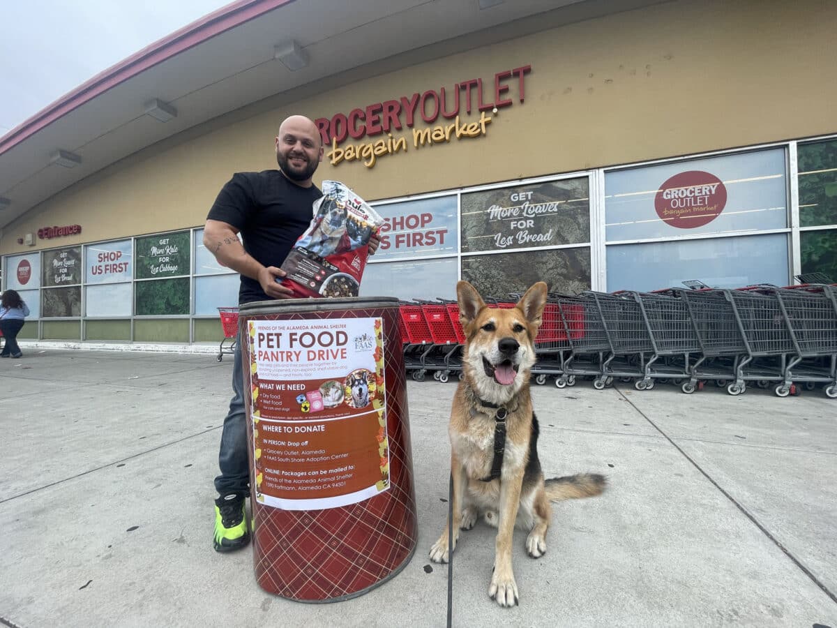 Alameda Post - Mouf the dog sits outside of Grocery Outlet next to a donation bin and a man smiling and holding a bag of dog food. 