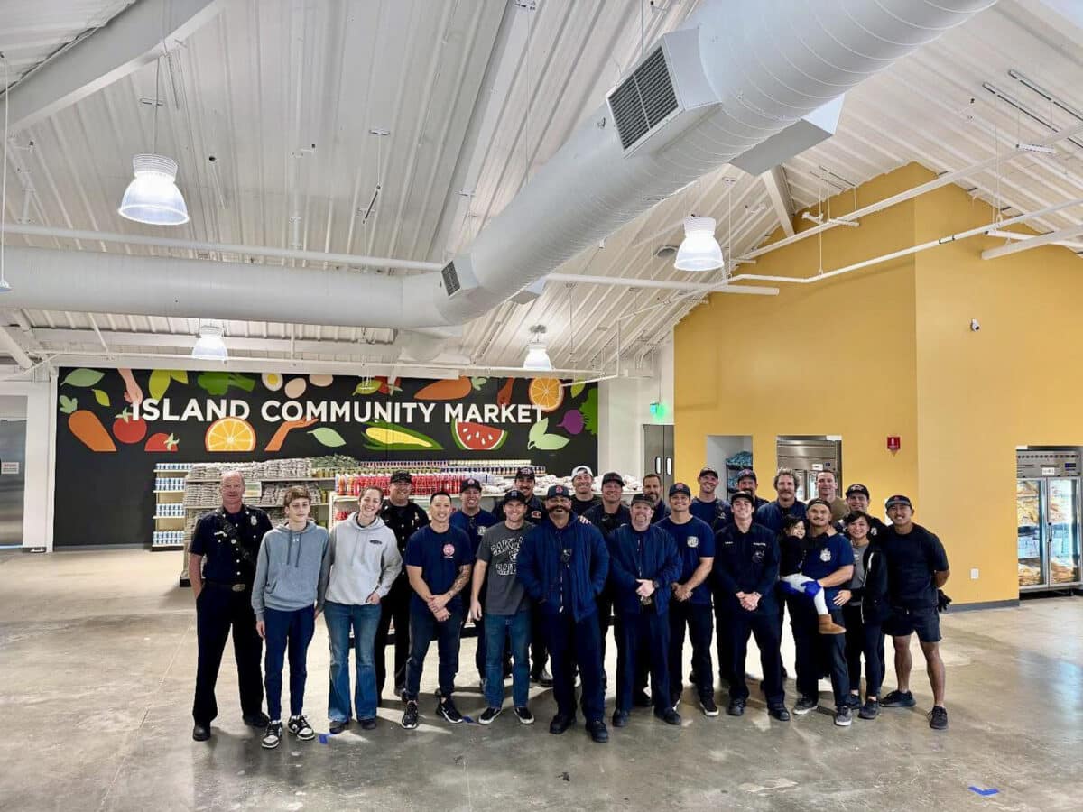 Alameda Post - Twenty four people including family members and kids stand and smile in the Island Community Market at the food bank. They are all firefighters, personnel, or family members. 