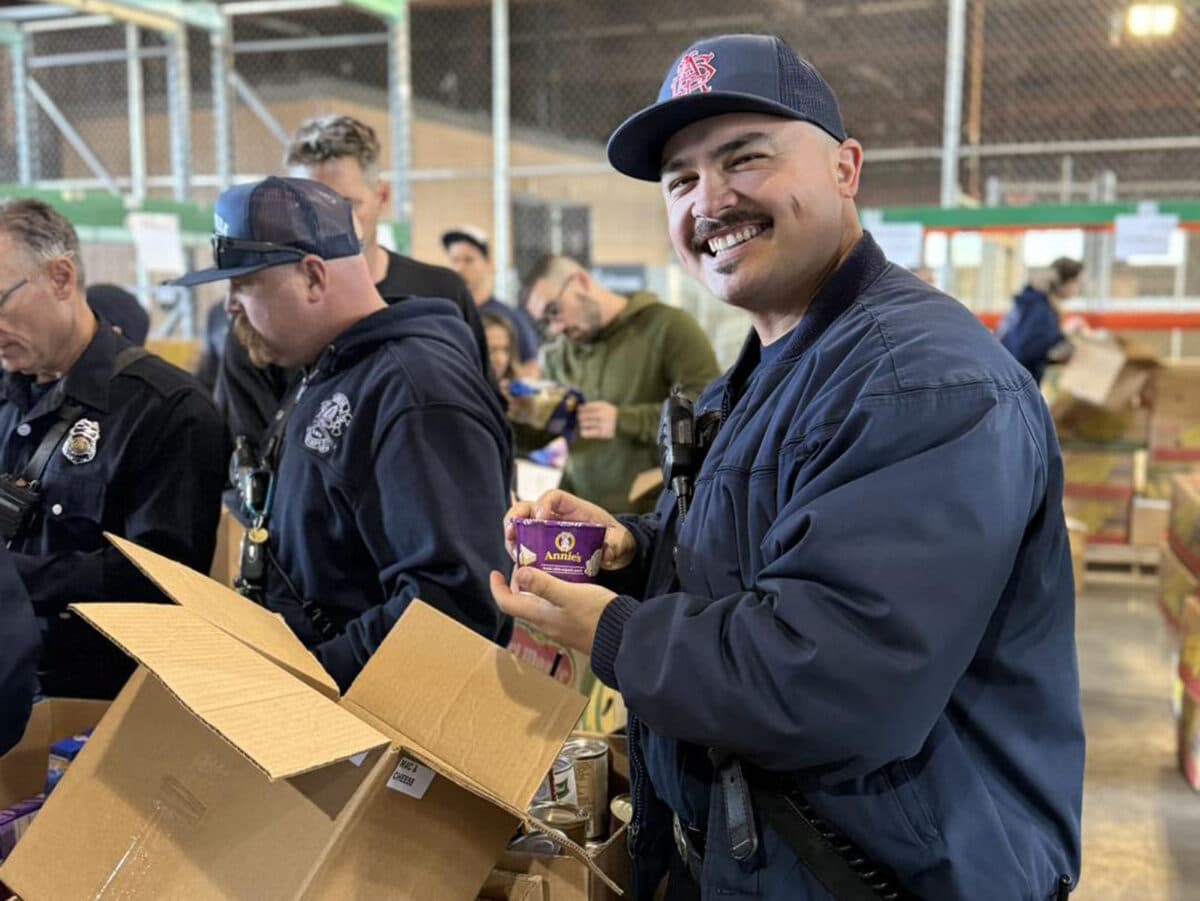 Alameda Post - An Alameda Fire Fighter smiles while holding a cup of mac and cheese. He is boxing items at the food bank.