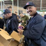 Alameda Post - An Alameda Fire Fighter smiles while holding a cup of mac and cheese. He is boxing items at the food bank.