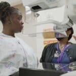 Alameda Post - A black female patient walks up to a mammogram machine while a black female technician or nurse explains how it works.