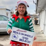 Alameda Post - A volunteer in an elf costume smiles and holds a sign that says "Alameda Meals on Wheels" with the phone number.