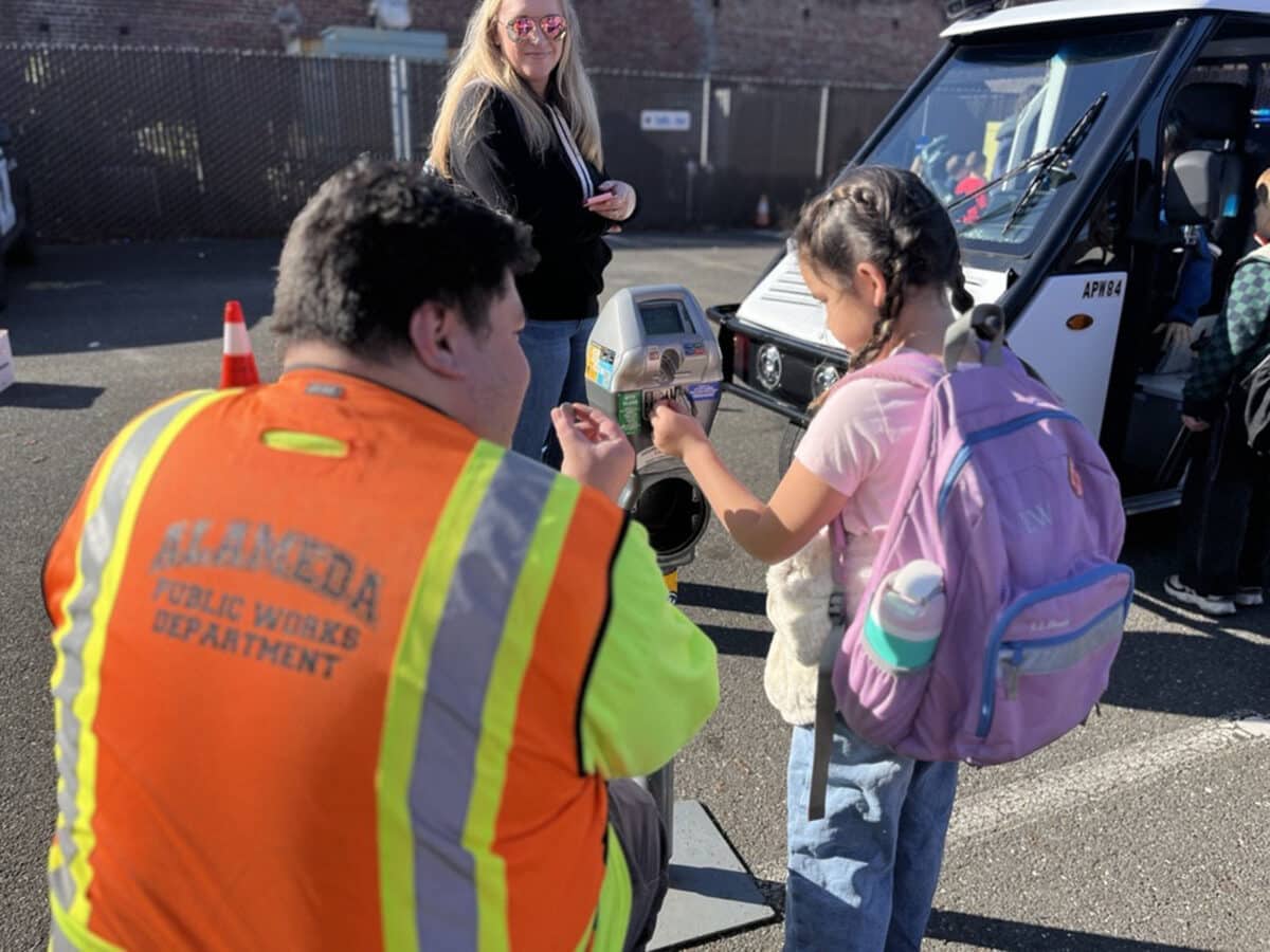 Alameda Post - An Alameda Public Works worker teaches a young school girl how to put quarters into a parking meter. 