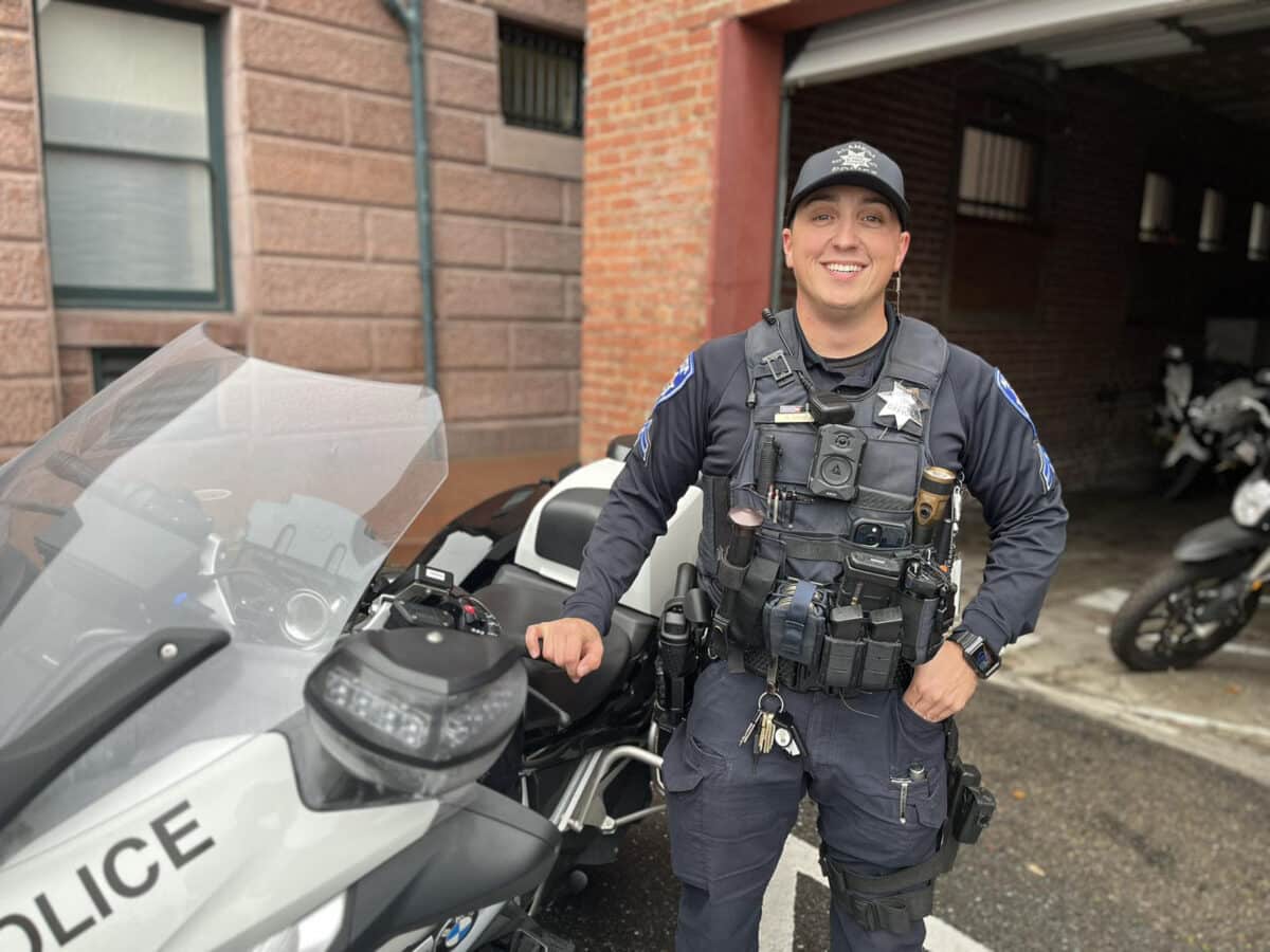 Alameda Post - Officer Sipes, APD's officer dedicated solely to traffic concerns, stands next to an APD motorcycle and smiles. 
